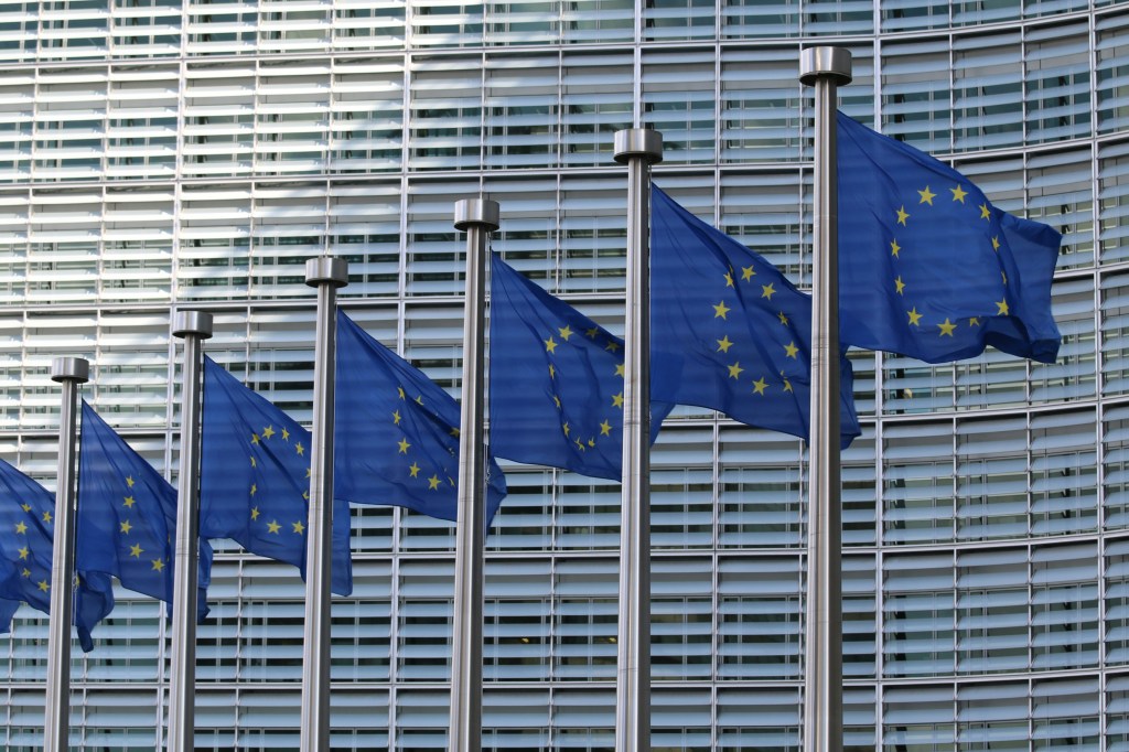 Multiple European Union flags flying in front of a modern glass building in Brussels.