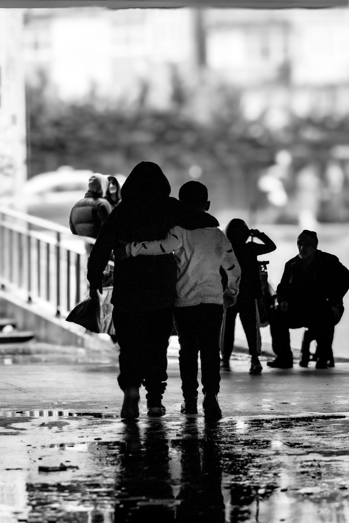 Black-and-white photograph of several people walking together through a covered urban passageway. In the foreground, an adult and a child walk side by side with their arms around each other. Other figures are visible in the background, including a seated person. The image evokes themes of displacement, support, and solidarity during uncertain times.