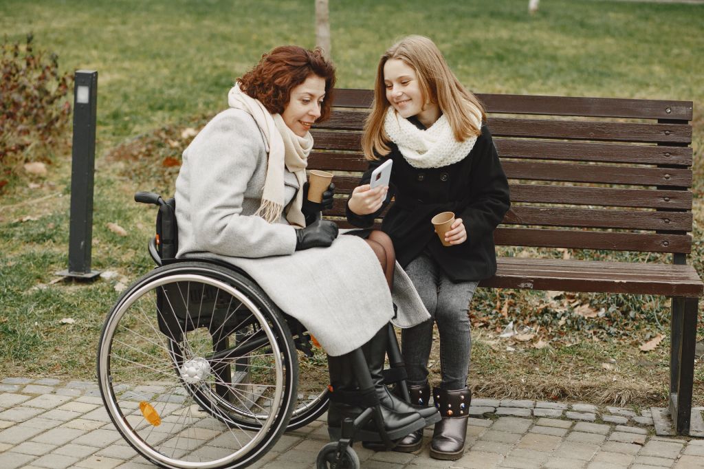 A woman using a wheelchair and a young girl sit together on a park bench, looking at a mobile phone and sharing a conversation. Photo credit by Gustavo Fring.