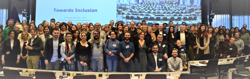 Group photo of participants at the "Towards Inclusion" conference held at the European Commission's Charlemagne Building in Brussels. Around 100 participants, including representatives of civil society organisations, EU institutions, service providers and persons with lived experience, are standing and sitting together in a conference room. A large screen behind the group displays the event title, "Towards Inclusion".