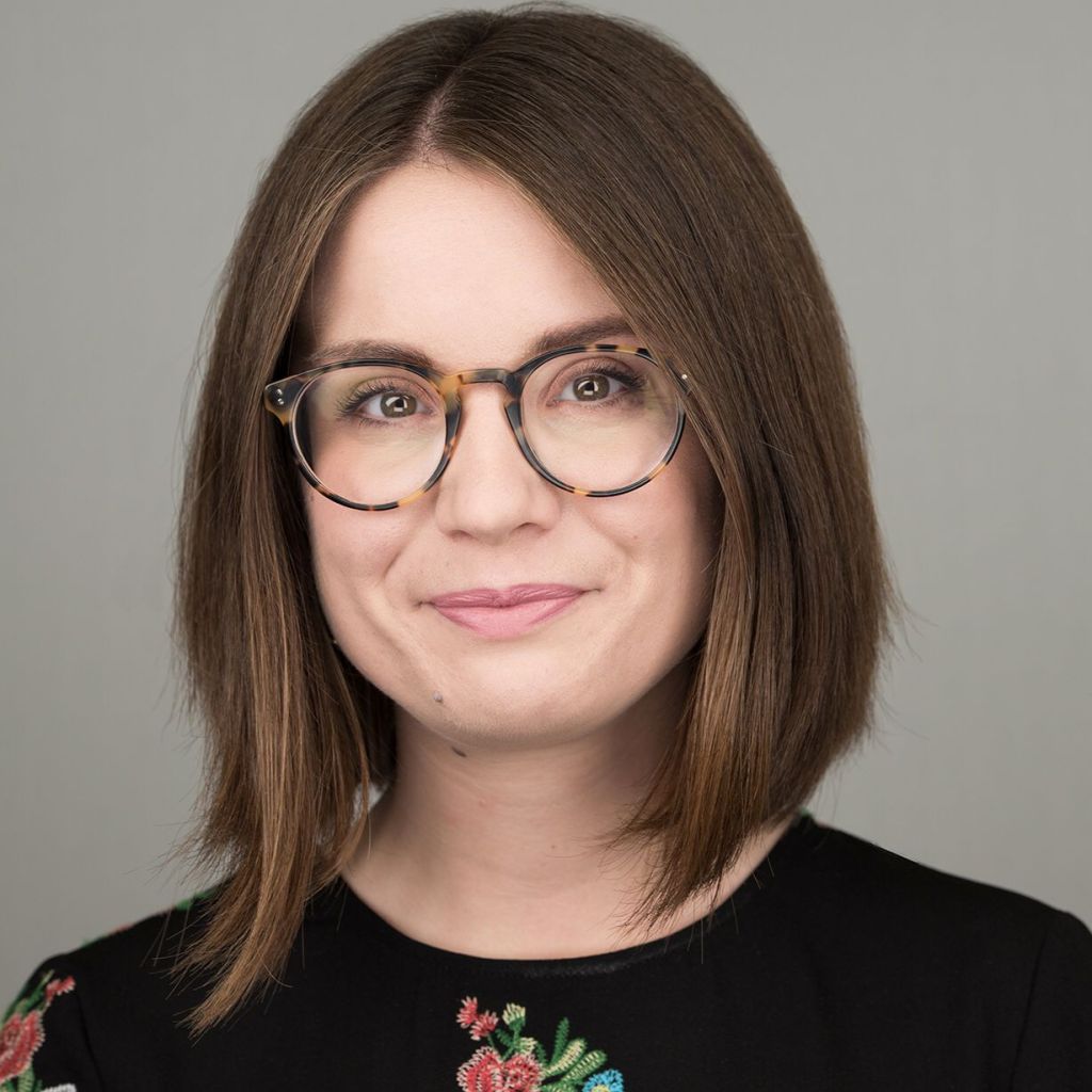 Professional headshot of Lucy Halton smiling at the camera. She has shoulder-length brown hair, wears round glasses and a black top with colourful floral embroidery, against a plain grey background.