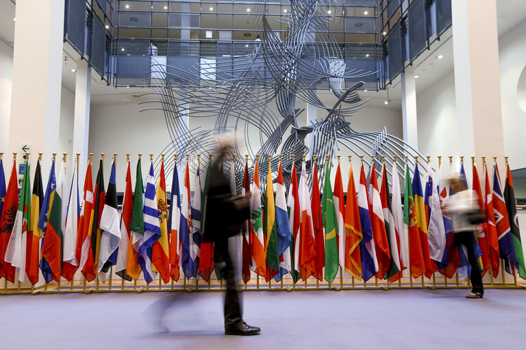 Interior of the European Parliament building with a row of European national flags and people walking past in the foreground.
