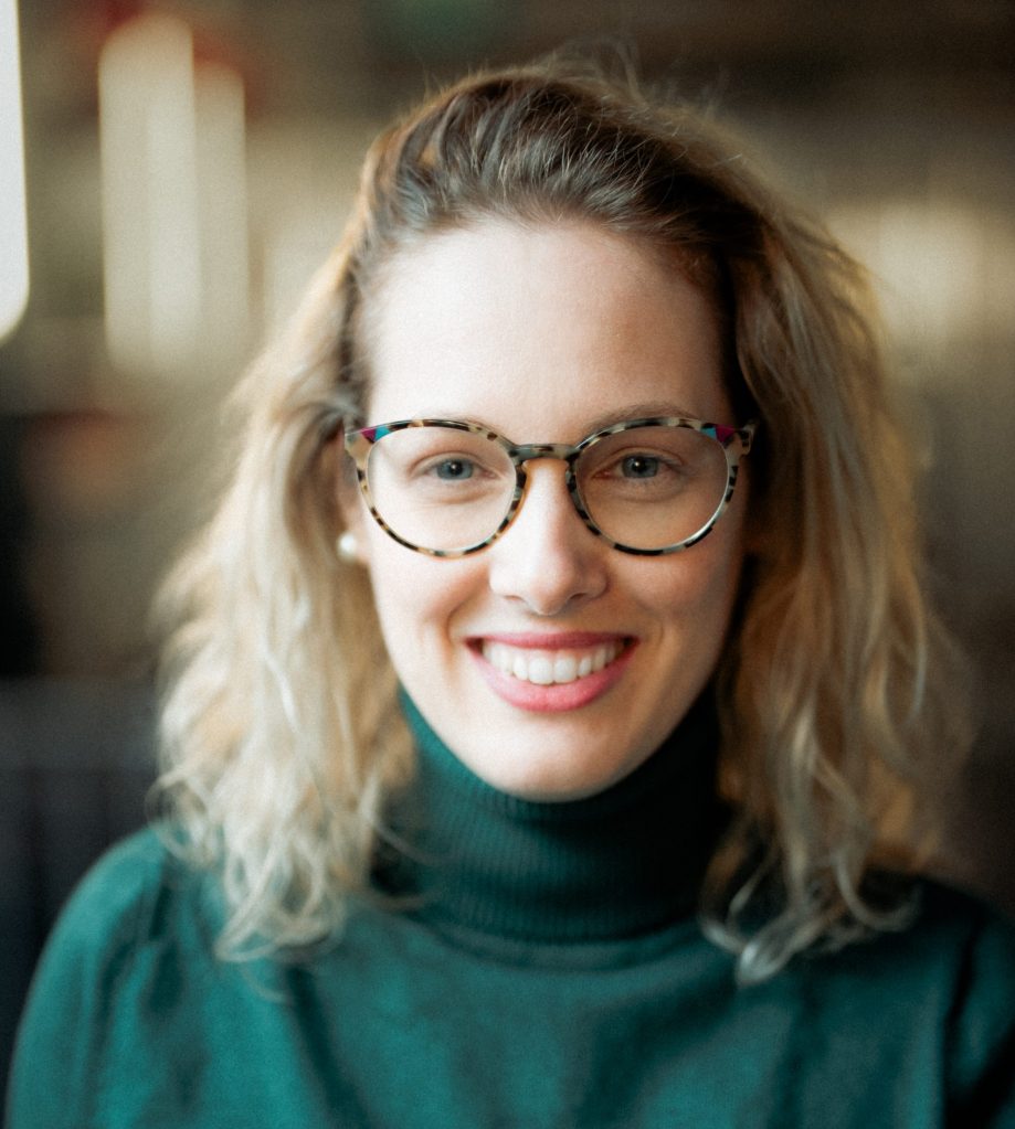 Portrait of Konstantina Leventi, smiling at the camera. She has shoulder-length blonde hair, wears patterned glasses, pearl earrings and a dark green turtleneck sweater. The background is softly blurred.